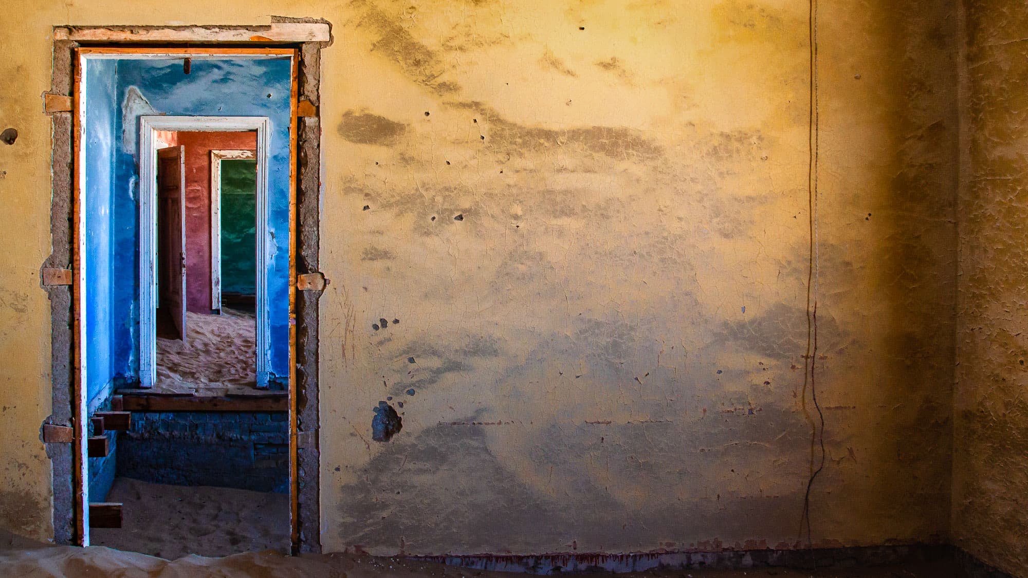 Kolmanskop erzählt Geschichten: Bunte Türrahmen führen durch verlassene Räume, während sich der Wüstensand die einstige Diamantenstadt bei Lüderitz langsam zurückholt.